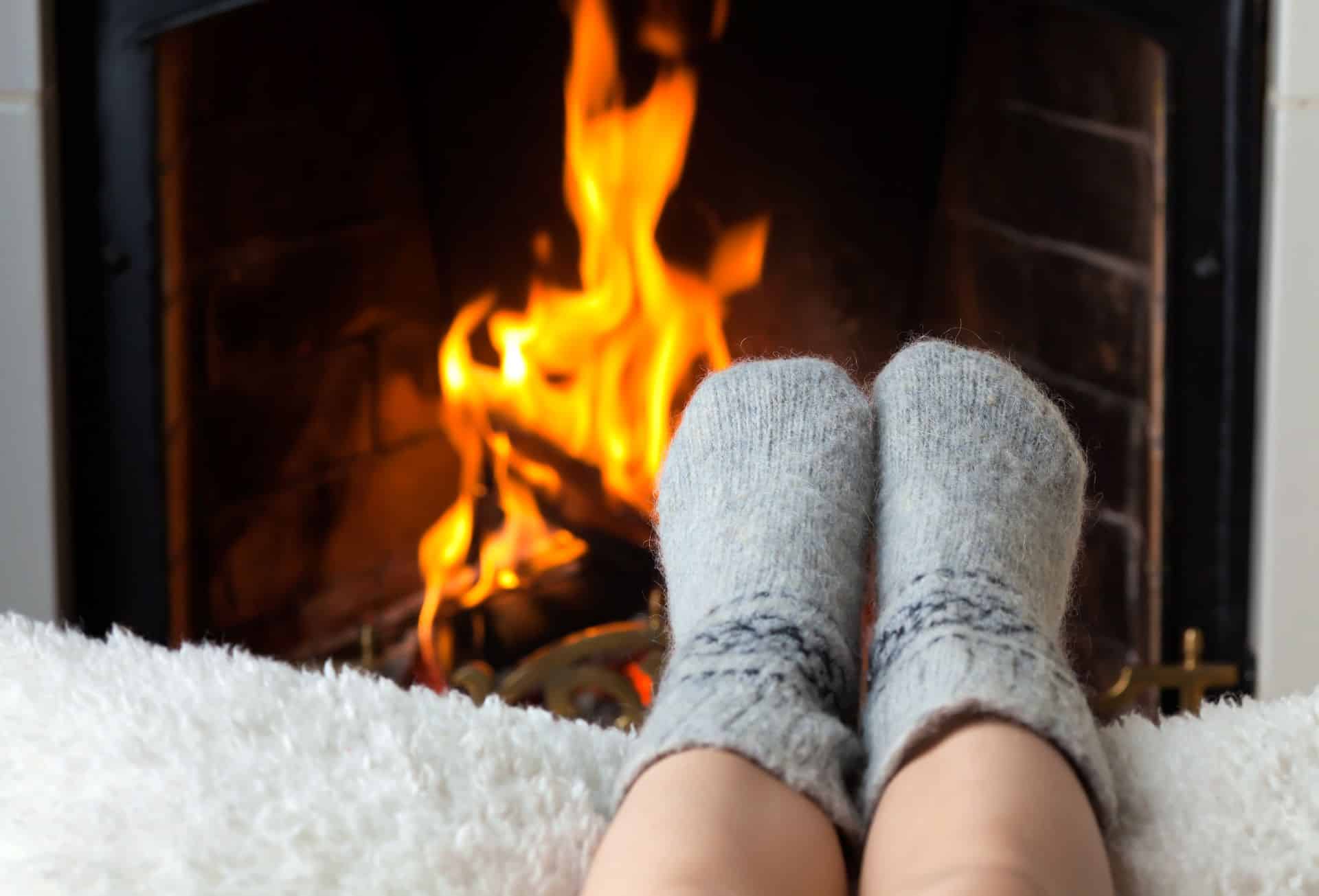 Zwei Füße in grauen Wollsocken, die vor einem lodernden Kaminfeuer auf einer weichen, weißen Decke ruhen. Die Flammen im Hintergrund sorgen für eine warme und behagliche Atmosphäre, während die dicken Socken Gemütlichkeit und Wärme vermitteln. Das Bild strahlt Ruhe und Entspannung aus, perfekt für eine gemütliche Zeit am Kamin.